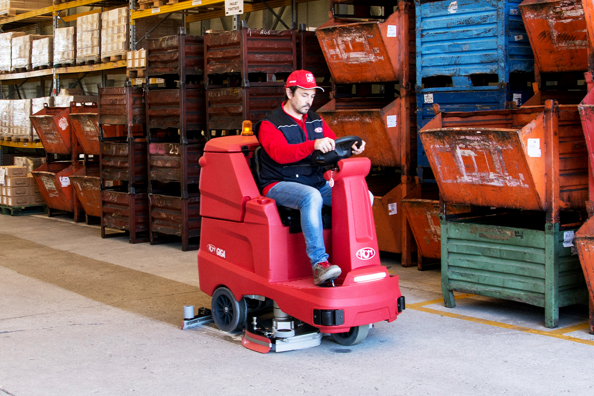 Ride-on floor scrubber cleaning an industrial workshop floor with storage bins in the background